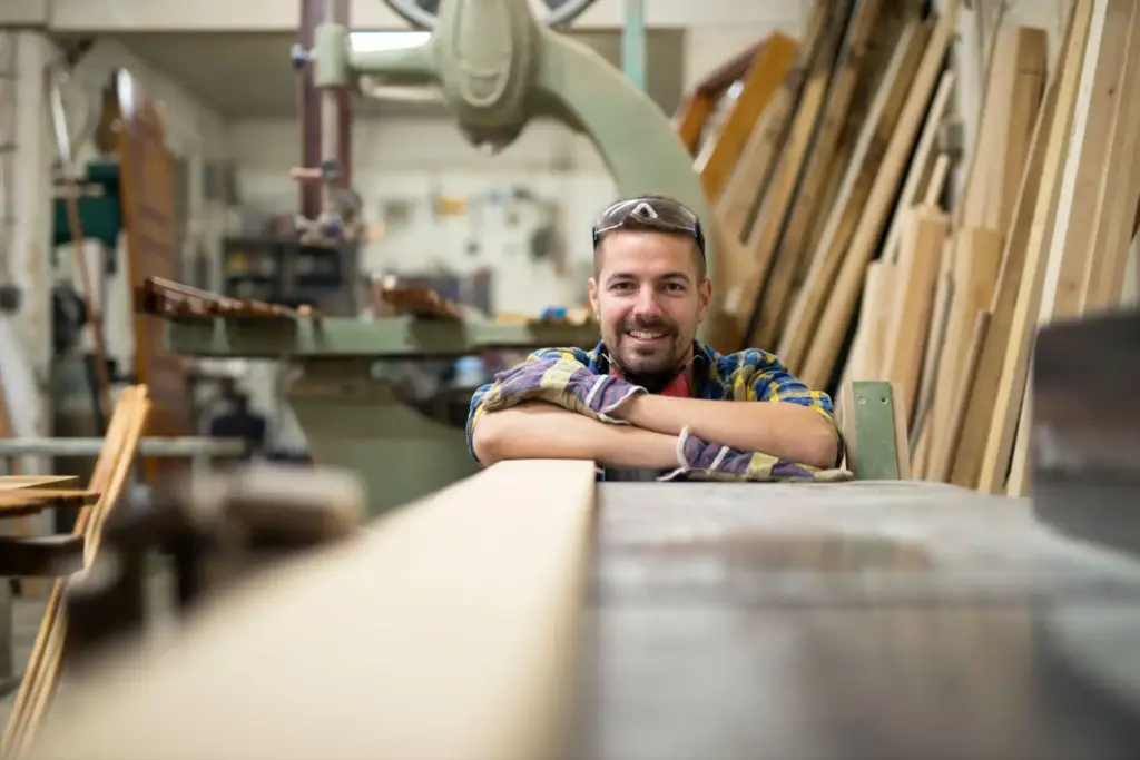 Um carpinteiro sorridente em uma oficina de madeira, com ferramentas e pranchas de madeira ao fundo para realizar projetos para marcenaria.