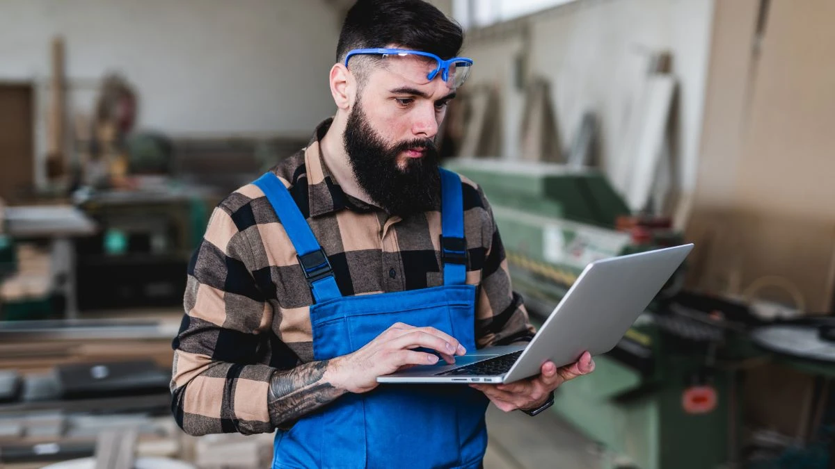 Homem com barba e óculos de proteção, usando laptop em um ambiente de trabalho entendendo como usar o Corte Certo. Ele veste um macacão azul e camisa xadrez, focado na tarefa.