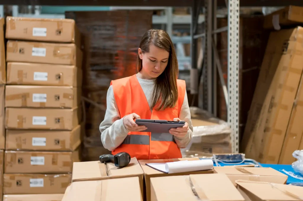 Mulher em armazém de produtos, usando colete de segurança laranja e inspecionando estoque com tablet, ao lado de caixas e prancheta, ambiente organizado e industrial.