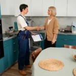 Profissional de entregas fazendo entrega a uma senhora na cozinha, com troca de sorrisos em ambiente doméstico moderno e bem decorado.